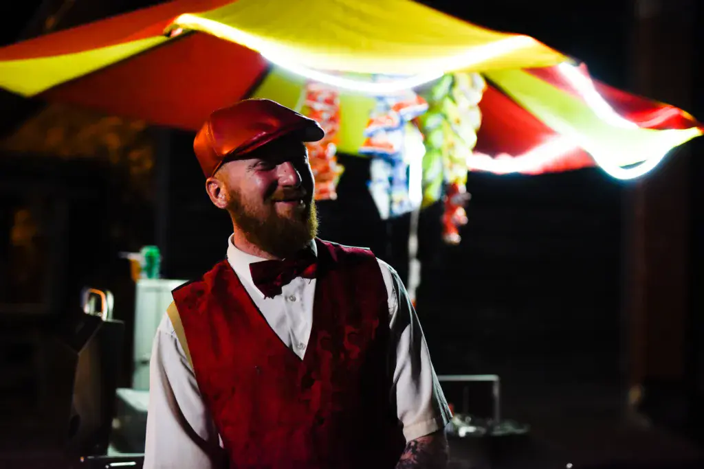 A performer in a red vest and cap stands on stage under colorful fabric awning at night.