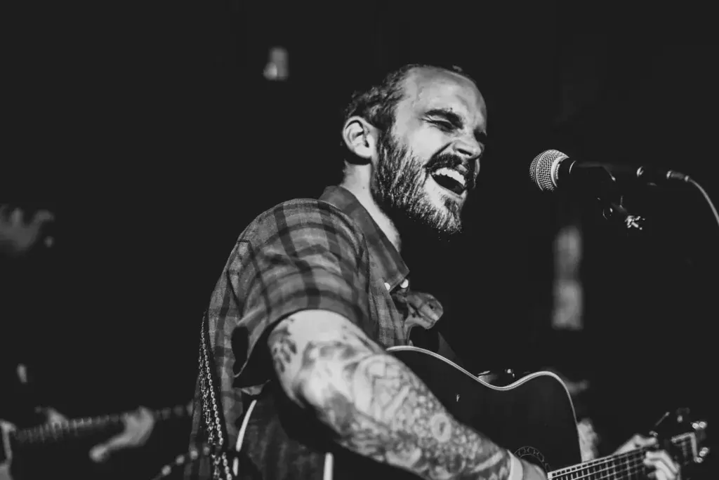 A bearded performer with a tattooed arm sings into a microphone while playing an acoustic guitar on stage.