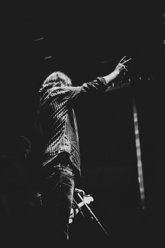 A performer on stage making a peace sign gesture while holding a guitar, photographed in black and white.