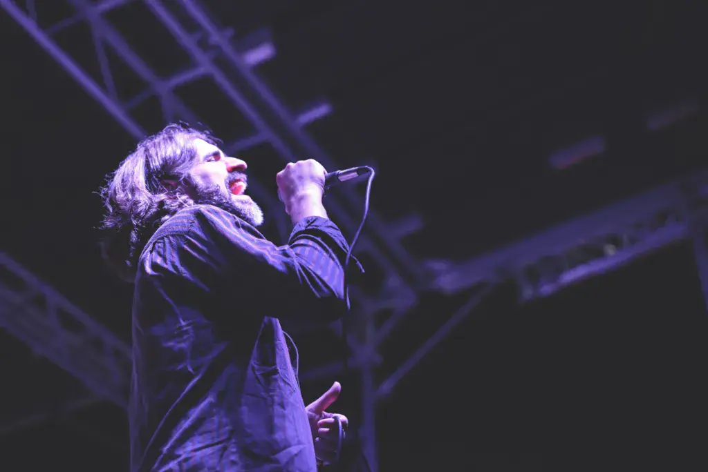 A performer with long hair singing into a microphone on stage under purple lighting.