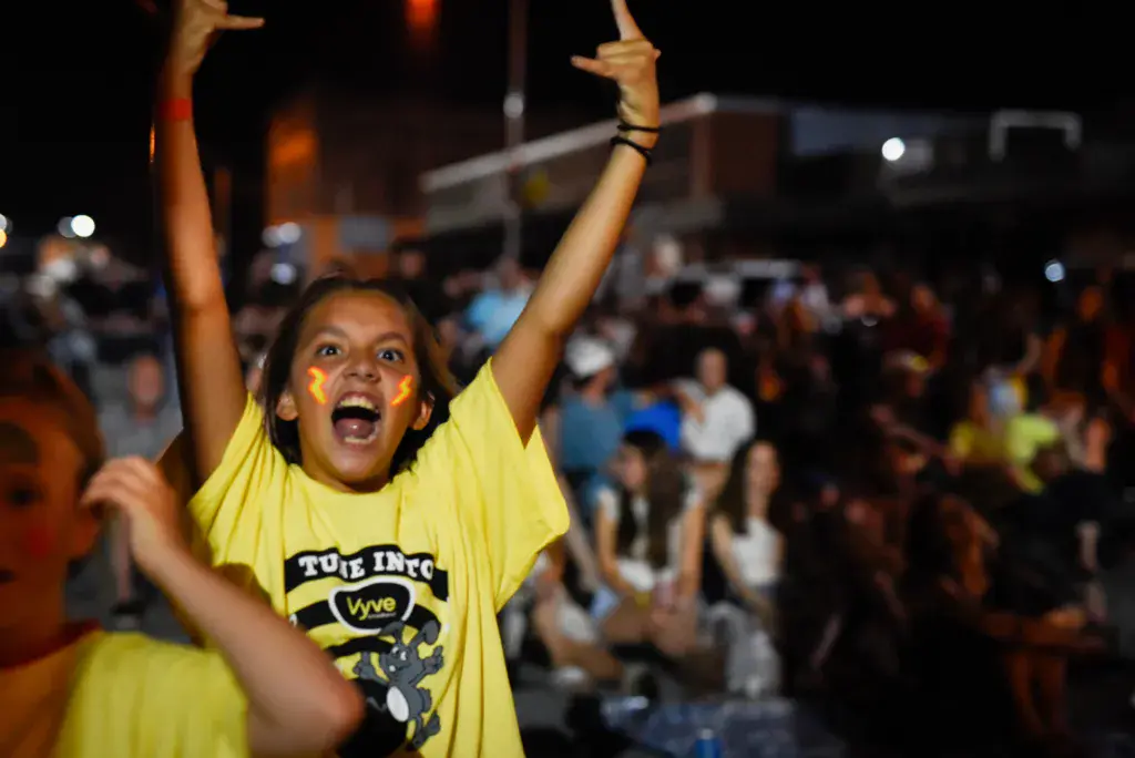 A young person in a yellow graphic t-shirt celebrates with arms raised at a nighttime outdoor music festival.