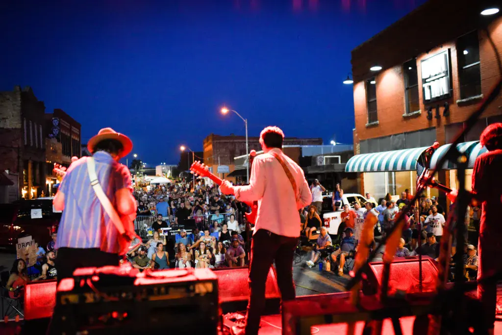 Band members perform on a red-lit stage as a large crowd watches during an evening outdoor festival.