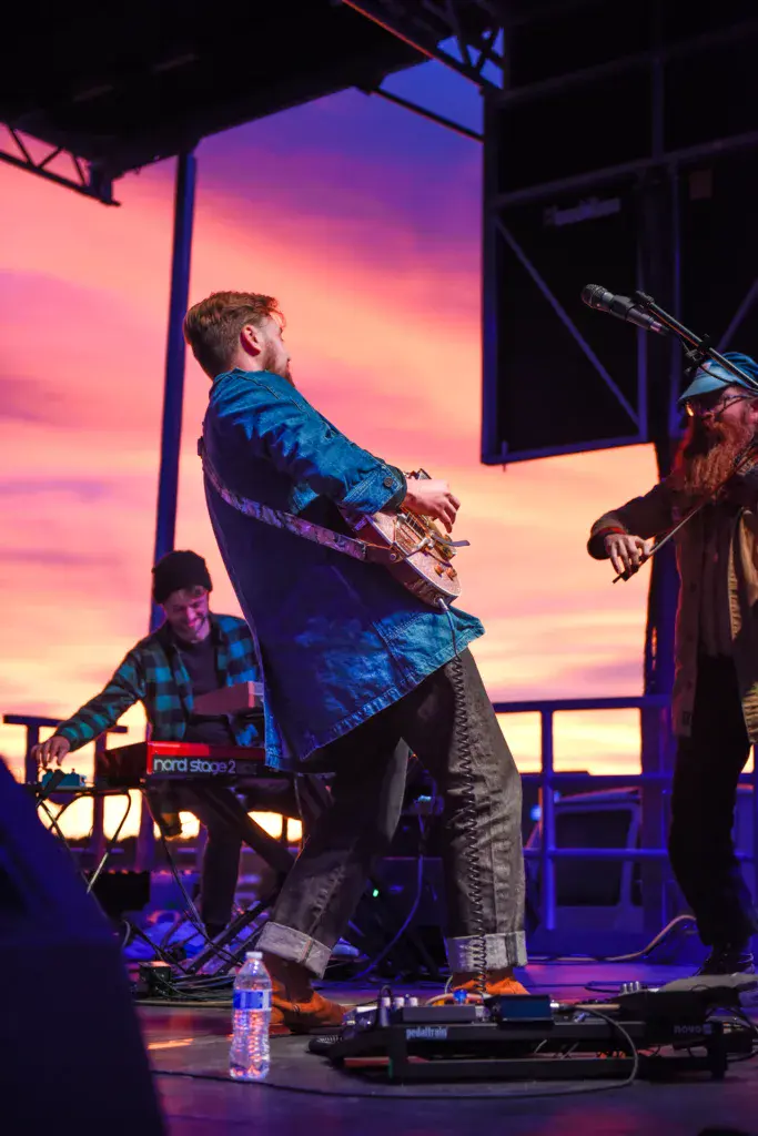 Musicians performing on stage with a vibrant pink and purple sunset backdrop during an outdoor music festival.