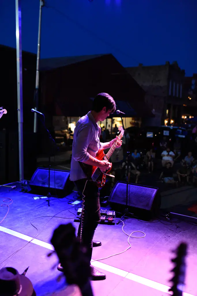 Guitarist performs on an outdoor stage with purple lighting while crowd watches at dusk.