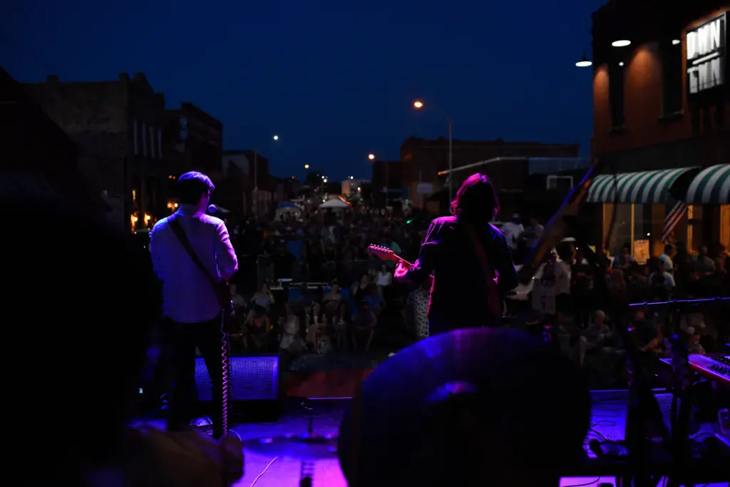 Musicians perform on stage at night with purple and blue stage lighting as the crowd watches in an urban outdoor venue.