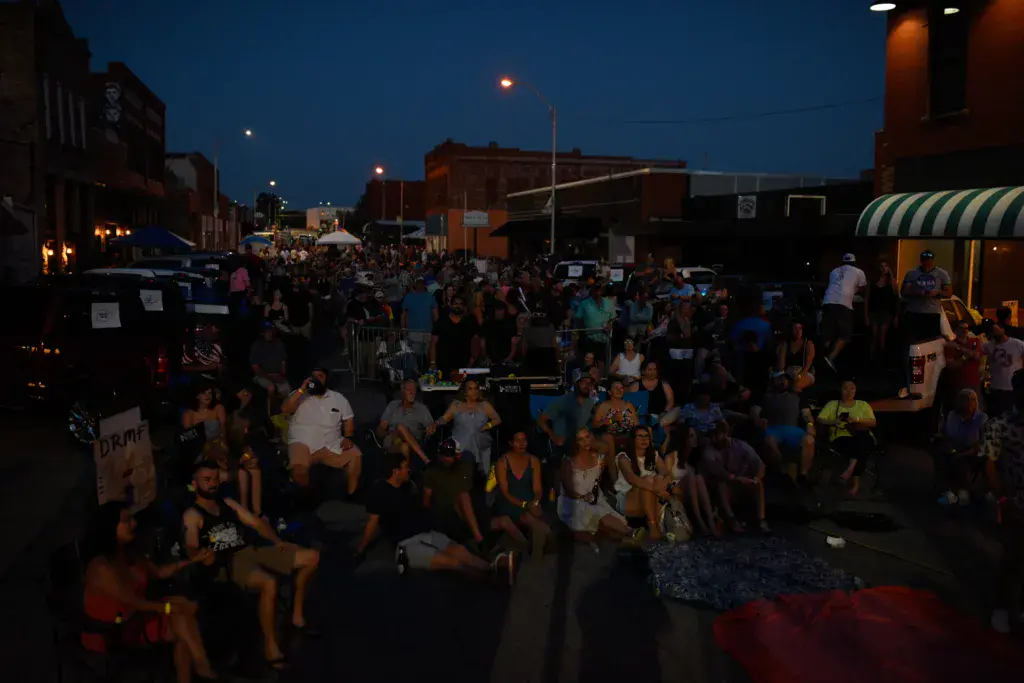 Large crowd gathered in an outdoor venue at dusk with illuminated buildings and vendor tents