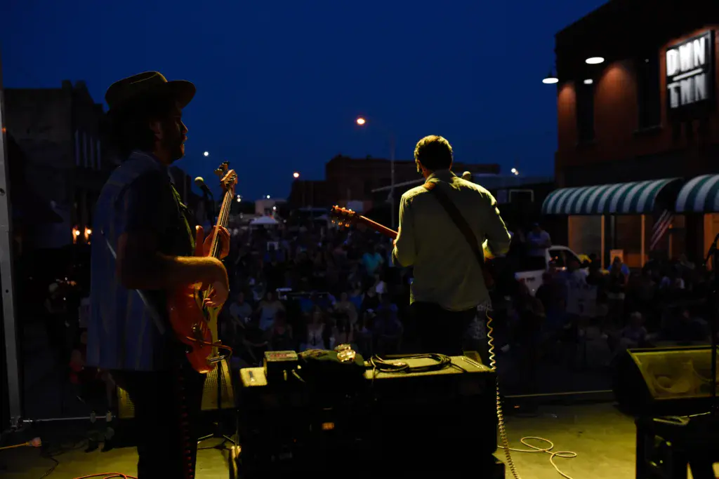 Musicians perform on an outdoor stage at dusk with a crowd watching in the background.