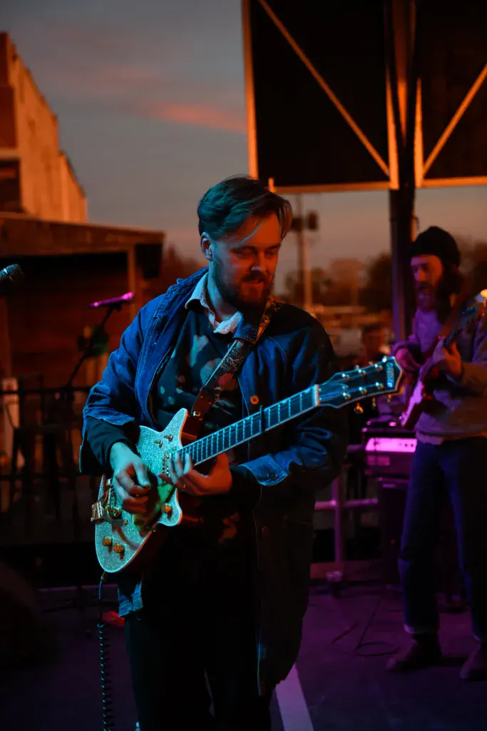 Guitarist performing on stage at dusk under orange-lit metal framework with band members visible in background.