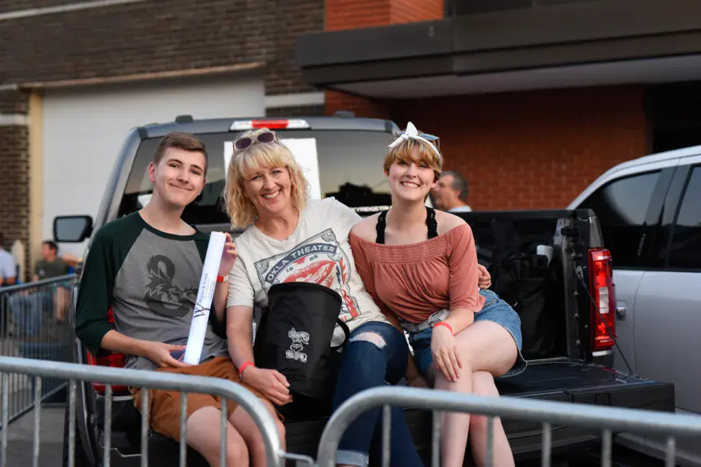 Three festival attendees smile while sitting on the tailgate of a parked vehicle in a venue parking area.