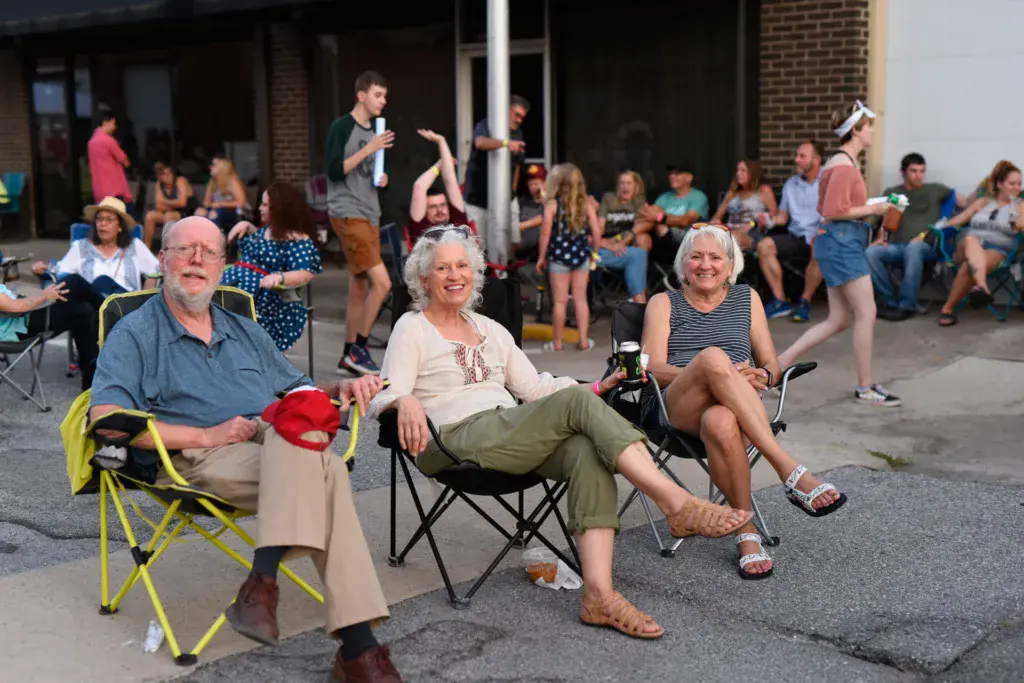 Festival attendees sitting and standing in a paved outdoor venue while a performer on stage addresses the crowd.