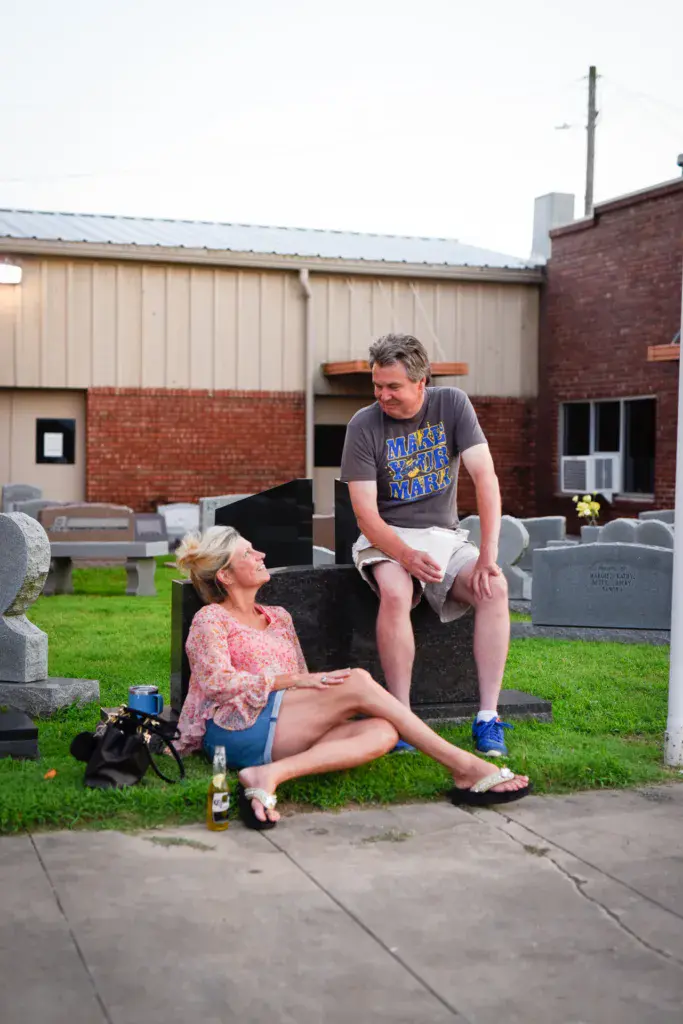 Two people sit on a gravestone at a cemetery in front of brick buildings.