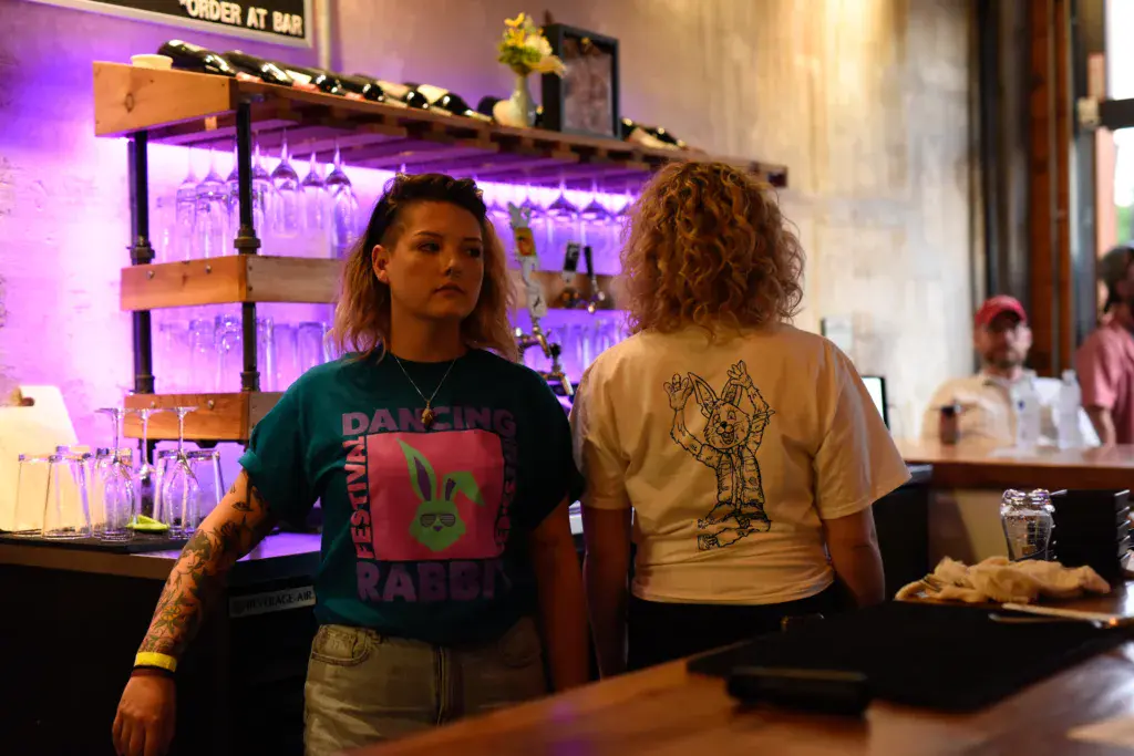Two festival attendees stand at a bar with purple-lit shelving displaying glasses in the background.