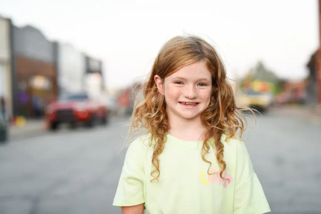 A young child with red hair wearing a light yellow shirt smiles at the camera at an outdoor festival venue.