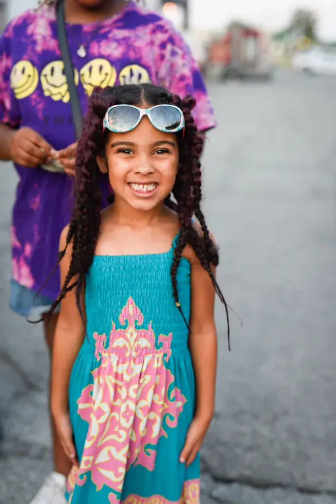 A smiling child wearing white sunglasses and a turquoise dress with colorful design stands at the festival.