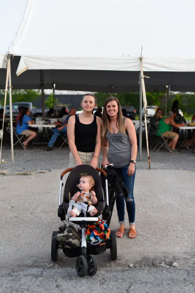 Two women stand with a stroller at the Dancing Rabbit Music Festival, with tent canopy and festivalgoers visible behind them.