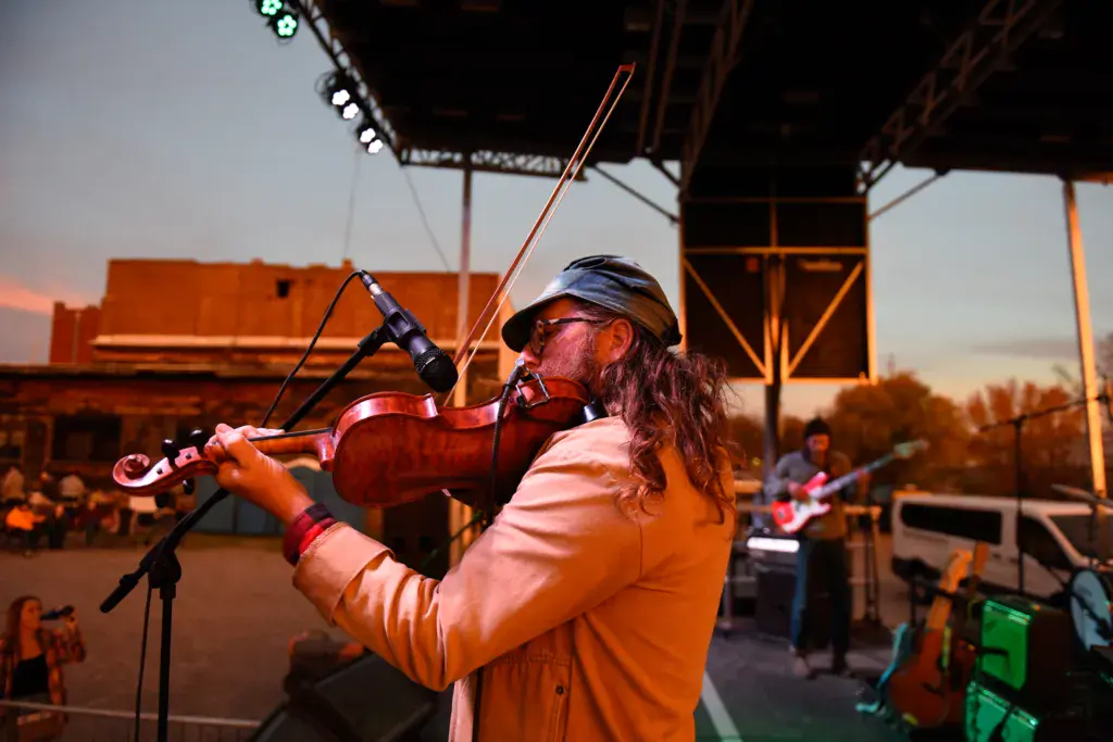 A performer plays violin into a microphone on stage during sunset at an outdoor music festival.