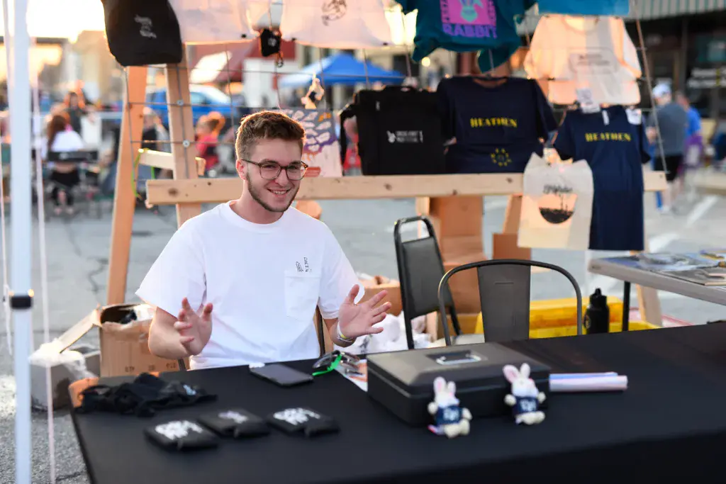 A person wearing glasses and a white t-shirt sits at a festival vendor booth displaying merchandise and promotional items.