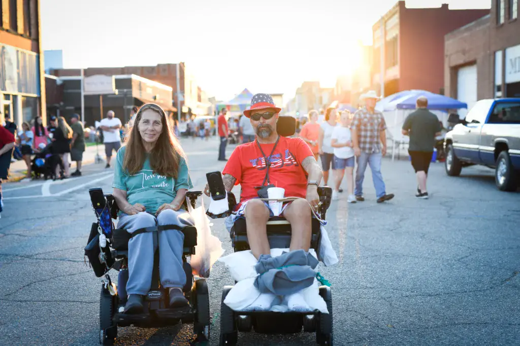 Two attendees in strollers smile at the camera during an outdoor festival on a sunny street lined with crowds and buildings.