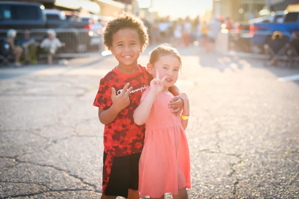 Two young children smiling at the camera on a festival grounds pathway during golden hour.