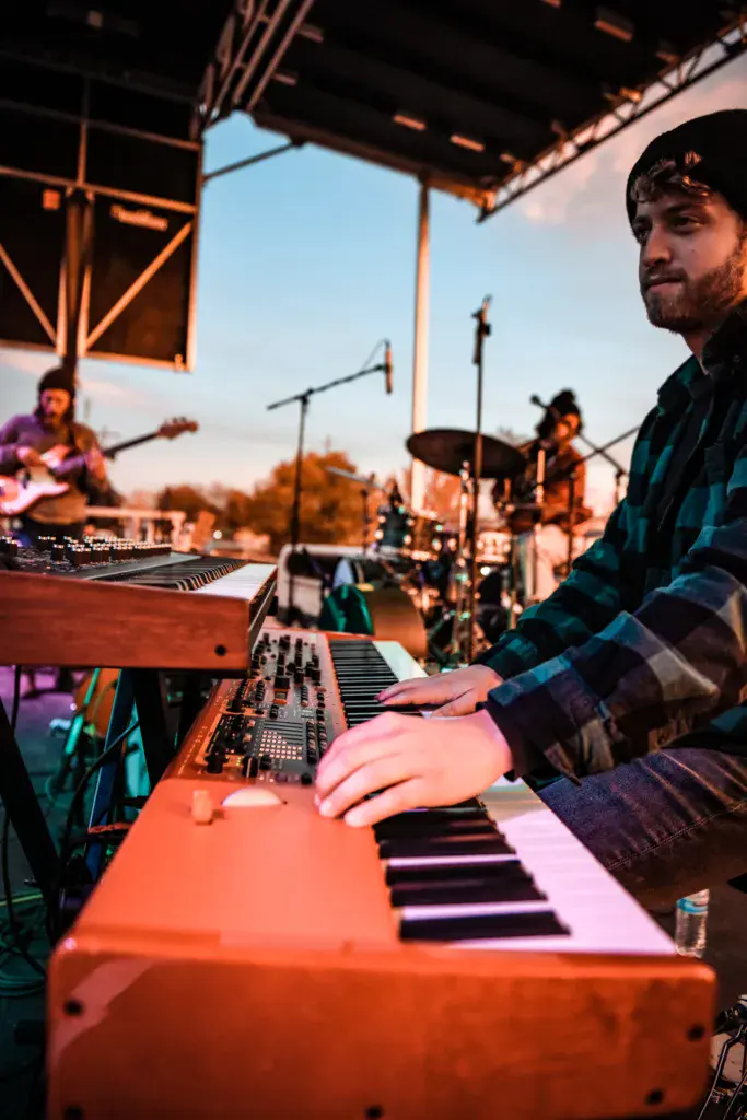 A keyboardist performs on stage at sunset during an outdoor music festival, with band members visible in the background.