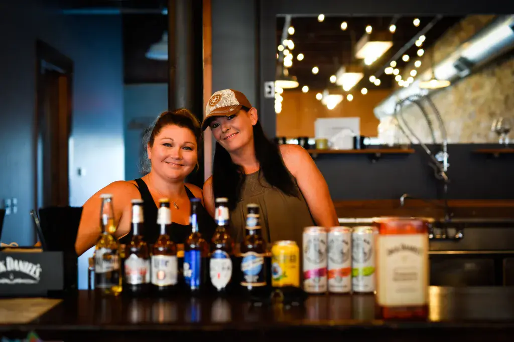 Two people smile at a bar counter with various alcoholic beverages displayed in front of them.