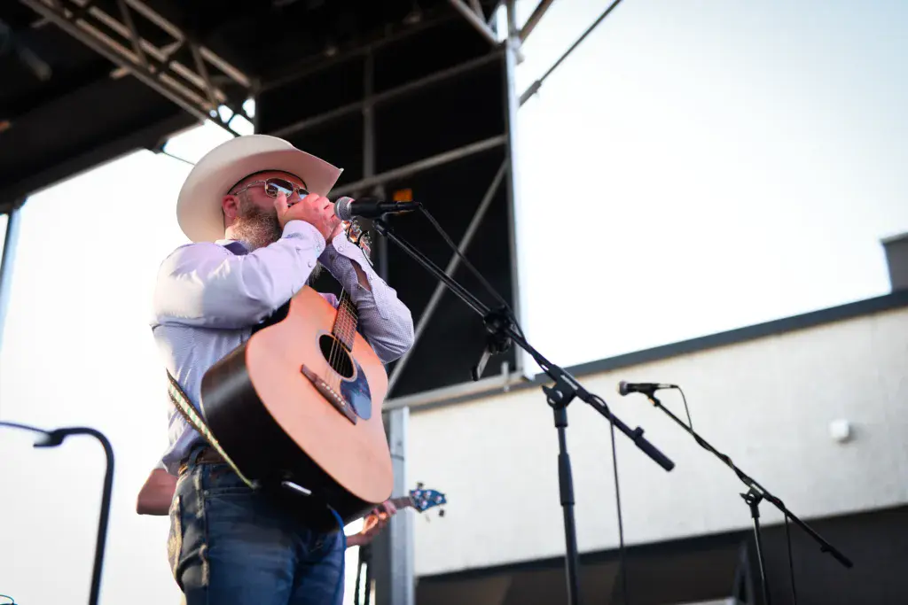 A performer wearing a white cowboy hat plays an acoustic guitar and sings into a microphone on stage.