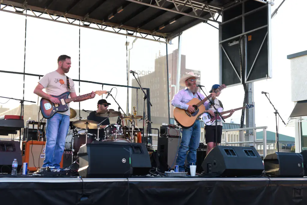 A four-piece band performs on an outdoor stage with a city skyline backdrop under a metal roof structure.