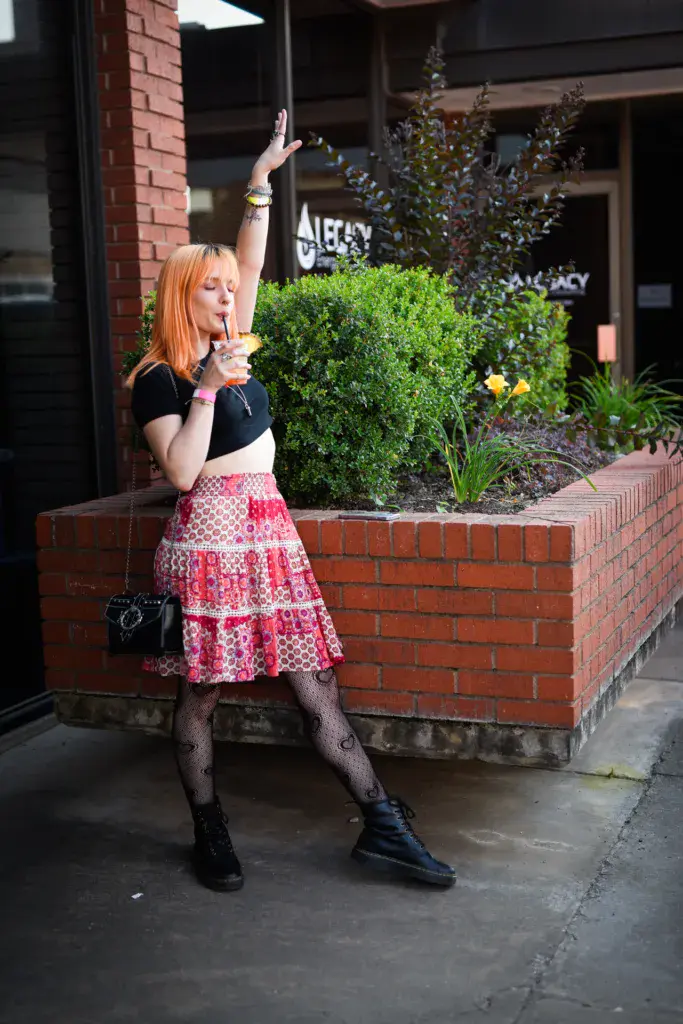 A performer with orange hair sings into a microphone outdoors against a brick venue backdrop.