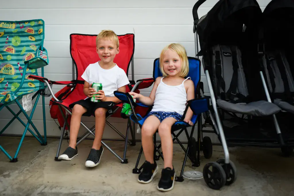 Two young children sitting in colorful lawn chairs holding drinks at an outdoor festival.