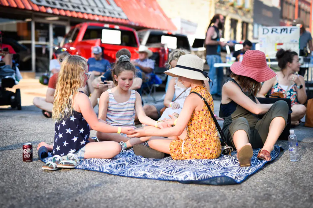 Young attendees sit together on a blanket at an outdoor festival venue with parked vehicles and other festival-goers in the background.