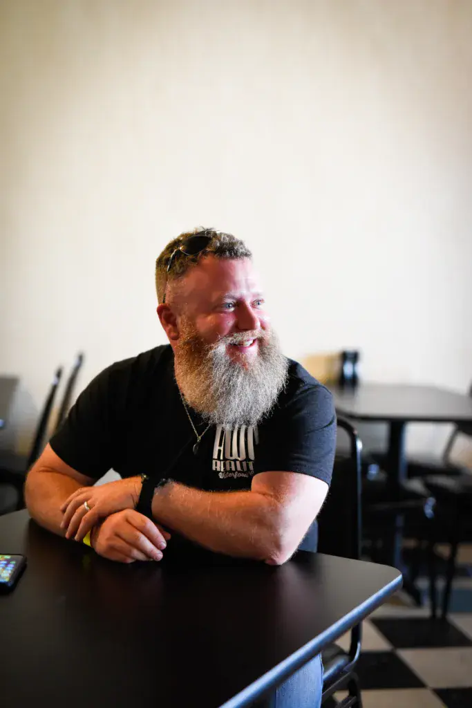 A bearded man in a black t-shirt sits at a table in an indoor venue, smiling while looking to the side.
