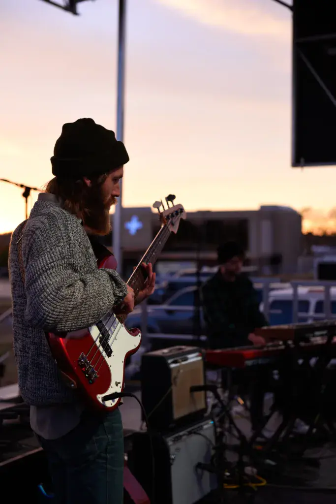 A musician in a beanie and gray cardigan performs with a red electric guitar during sunset at an outdoor festival.