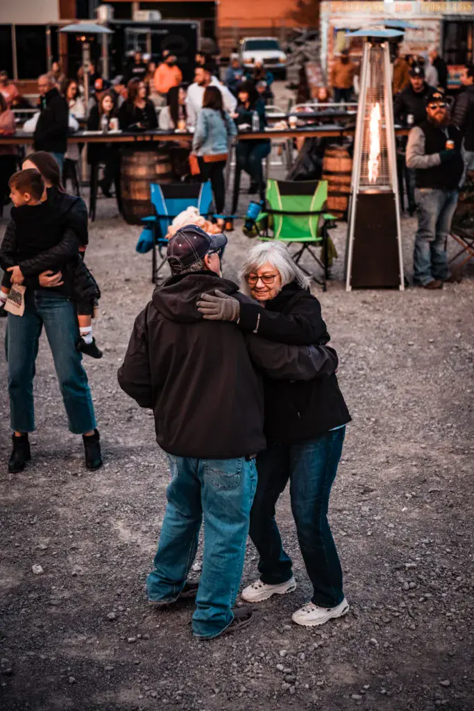 Two people embrace at an outdoor festival venue with crowds and camping chairs visible in the background.