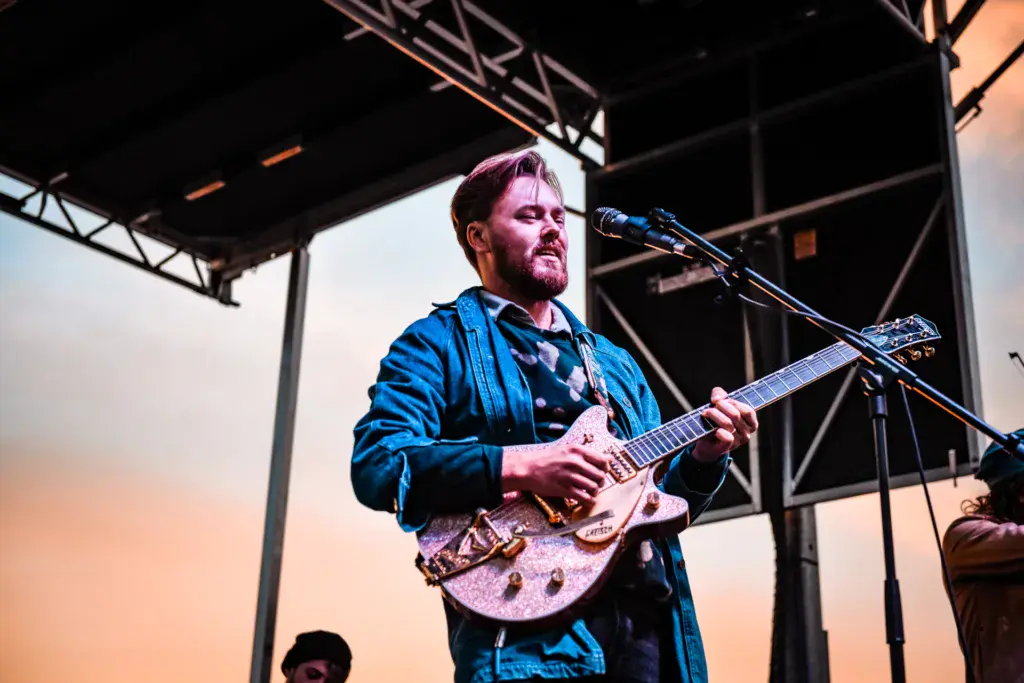 Guitarist performs at sunset on stage at Dancing Rabbit Music Festival under a metal canopy structure.