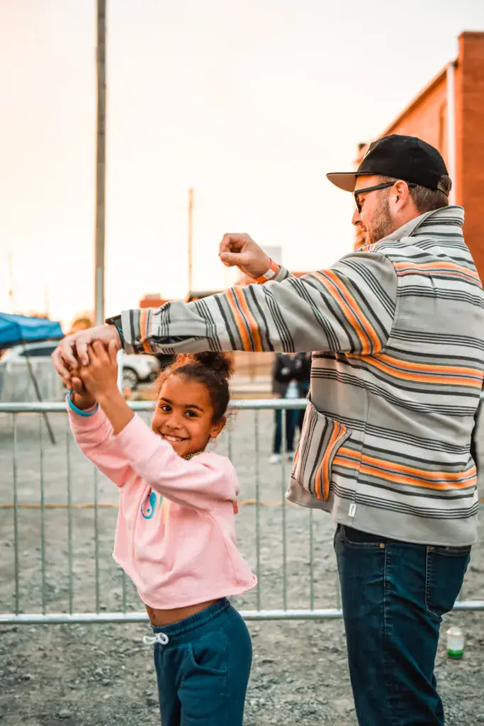 An adult and child dance together at the festival with a metal barrier and venue structures in the background.