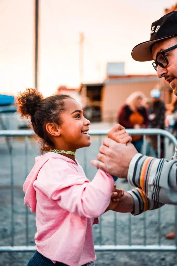 A young child in a pink shirt holds hands with an adult wearing a black cap at a festival venue with barriers and spectators in the background.