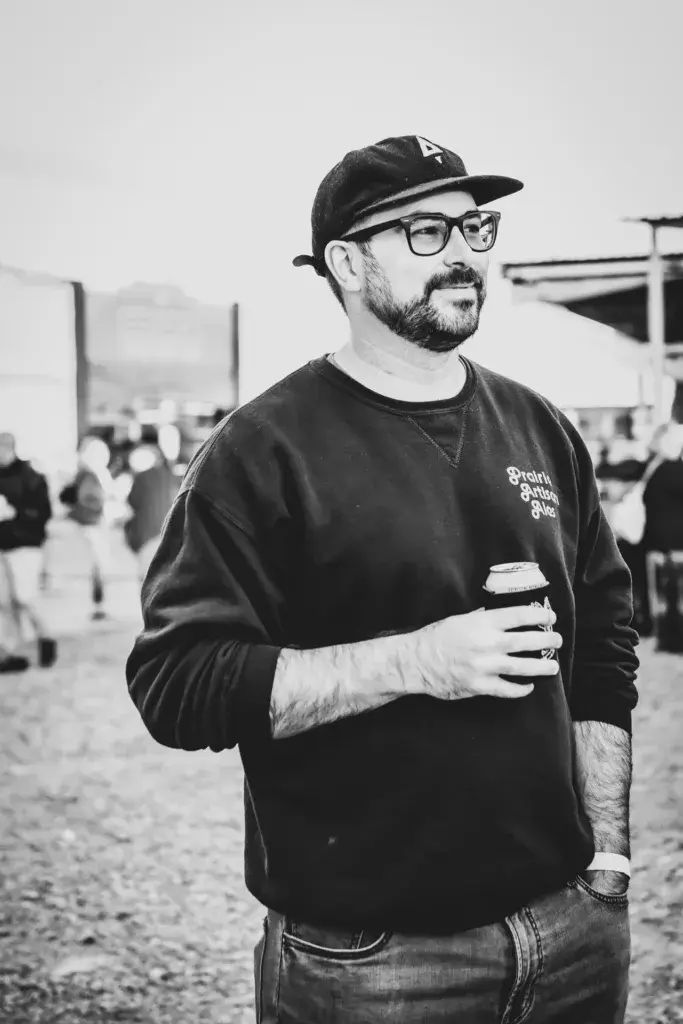 A man wearing glasses and a baseball cap holds a beverage at an outdoor festival with crowds visible in the background.