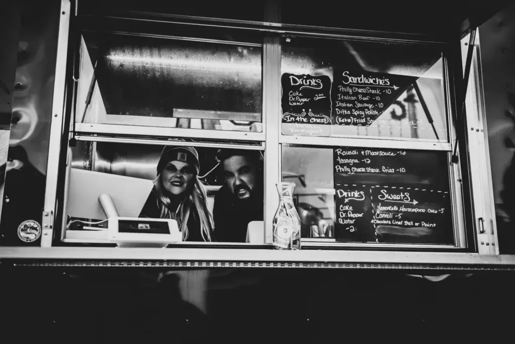 Two workers smile from inside a food truck window displaying menu boards with drinks and sandwich options.