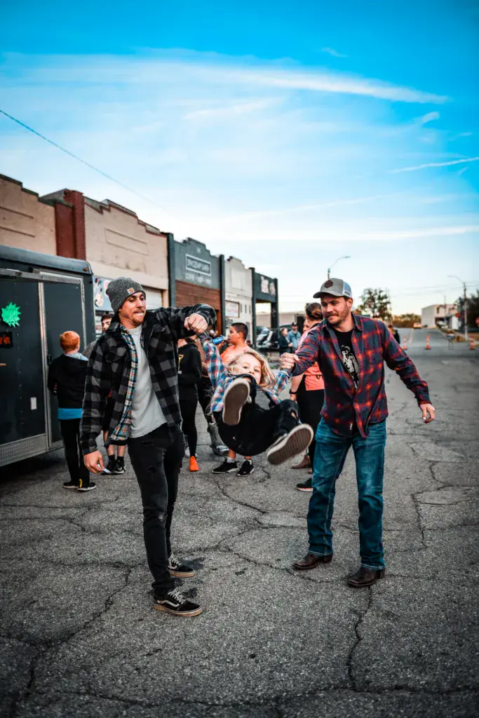 Festival attendees walking and interacting in a parking lot venue area with food trucks and buildings in the background.