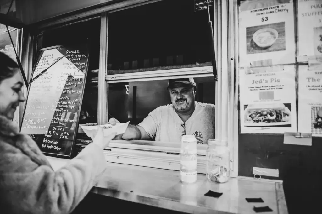 A vendor at a festival food stand serves a customer from a window counter in black and white.