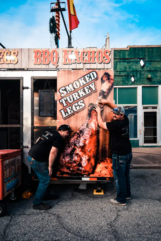 Two workers operate a food truck serving BBQ nachos and smoked turkey legs at an outdoor festival.