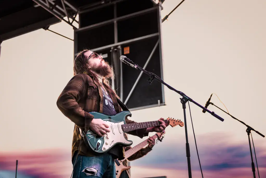 A musician with long hair and glasses plays electric guitar at a microphone on stage against a purple sunset sky.