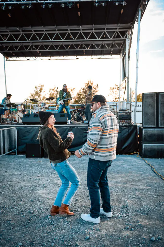 Two people interact on the festival grounds in front of the main stage with musicians performing in the background.