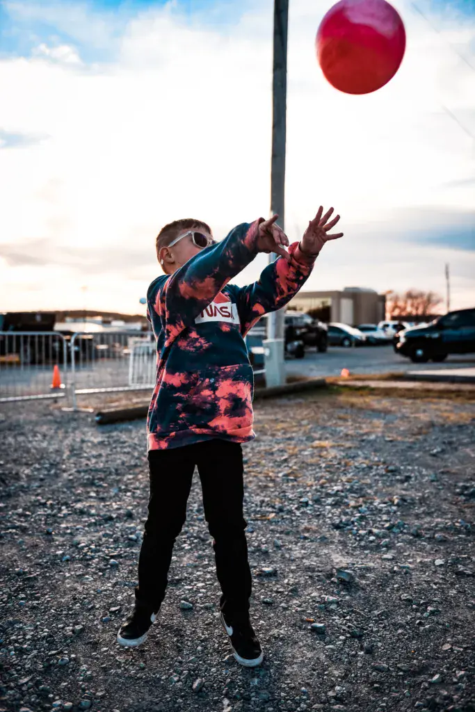 A young person in a tie-dye hoodie and sunglasses reaches up to catch a red ball against a cloudy sky.