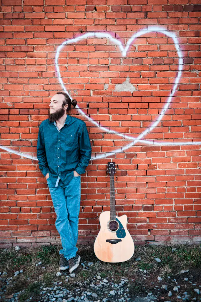 A musician with a beard stands against a red brick wall with a heart outline, holding an acoustic guitar.