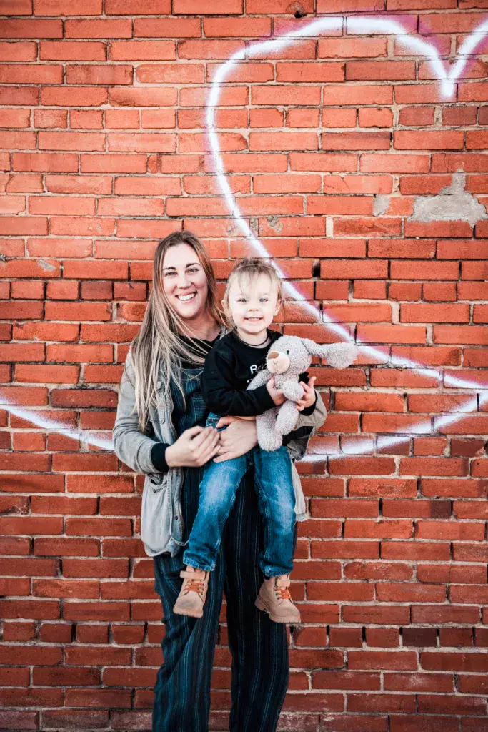 An adult and young child pose together holding a stuffed animal in front of a red brick wall with white graffiti art.