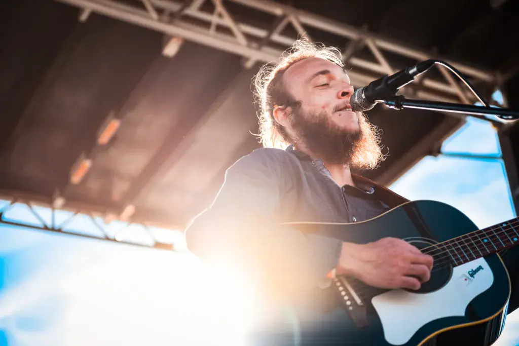 A bearded performer with long hair sings into a microphone while playing an acoustic guitar on an outdoor festival stage.