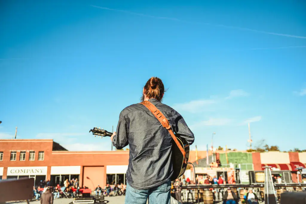 Performer with guitar and orange strap stands on stage facing a crowd at an outdoor festival venue.