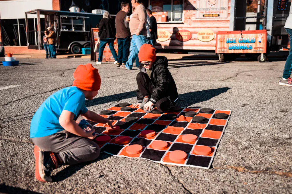 Two children wearing orange beanies play a large outdoor checkers game at a festival venue.
