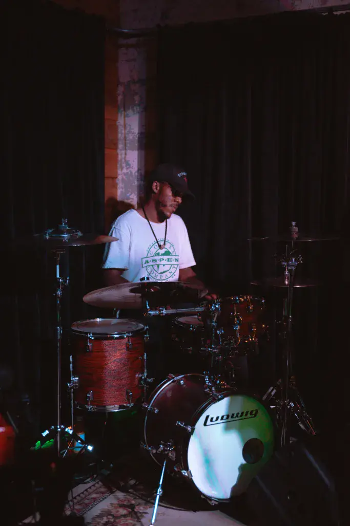 A drummer performs on stage at an indoor venue, playing a red drum kit with dramatic lighting.
